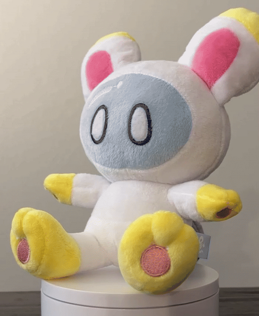 A plush toy character with a white body, light blue face, yellow feet and hands, and pink inner ears, sitting on a white cylindrical pedestal against a plain indoor background.